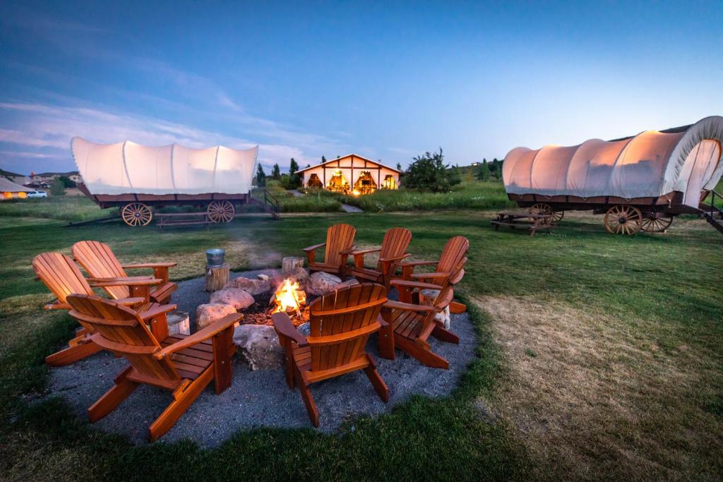 Kring van houten stoelen rond een kampvuur bij schemering, met verlichte huifkarren en een lodge op de achtergrond