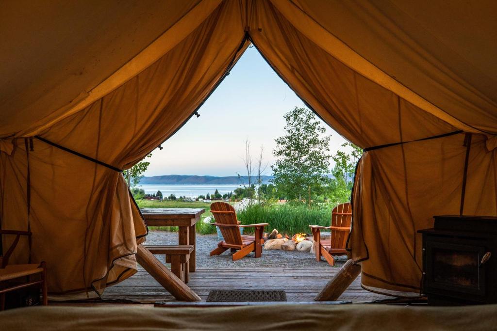 Blik vanuit de opening van een glampingtent naar buiten, met een houten tafel, stoelen bij een vuurkorf en het meer op de achtergrond