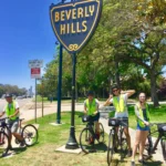 Vier fietsers met gele veiligheidshesjes en helmen poseren lachend bij het bekende Beverly Hills straatbord tijdens een fietstour