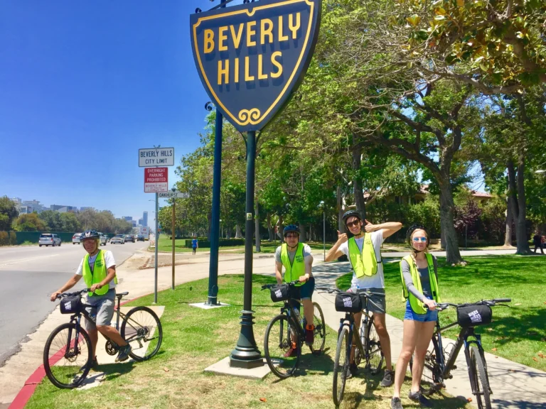 Vier fietsers met gele veiligheidshesjes en helmen poseren lachend bij het bekende Beverly Hills straatbord tijdens een fietstour