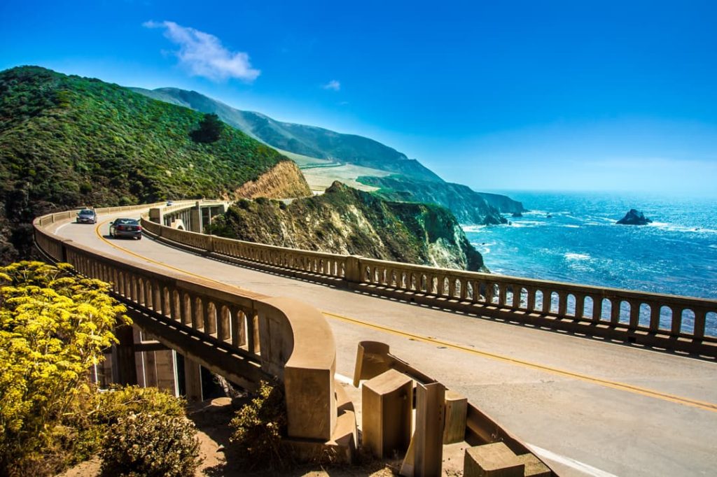 Gebogen betonnen brug waarover auto's rijden, met steile groene heuvels en de blauwe oceaan op de achtergrond bij Big Sur