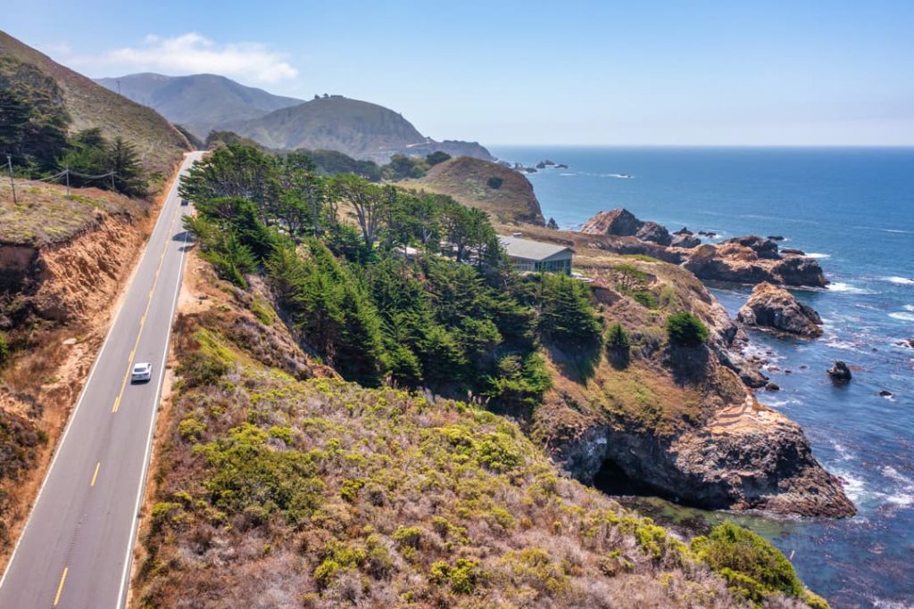 Een witte auto rijdt over een kronkelende kustweg omzoomd door bomen met uitzicht op een rotsachtige kust en helderblauwe oceaan bij Big Sur