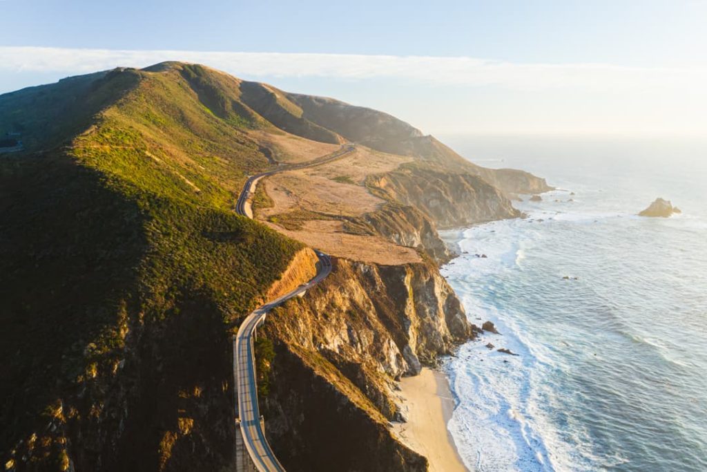 Luchtfoto van een asfaltweg die over een groene, glooiende kaap loopt met een zandstrand en de oceaan aan weerszijden bij Big Sur