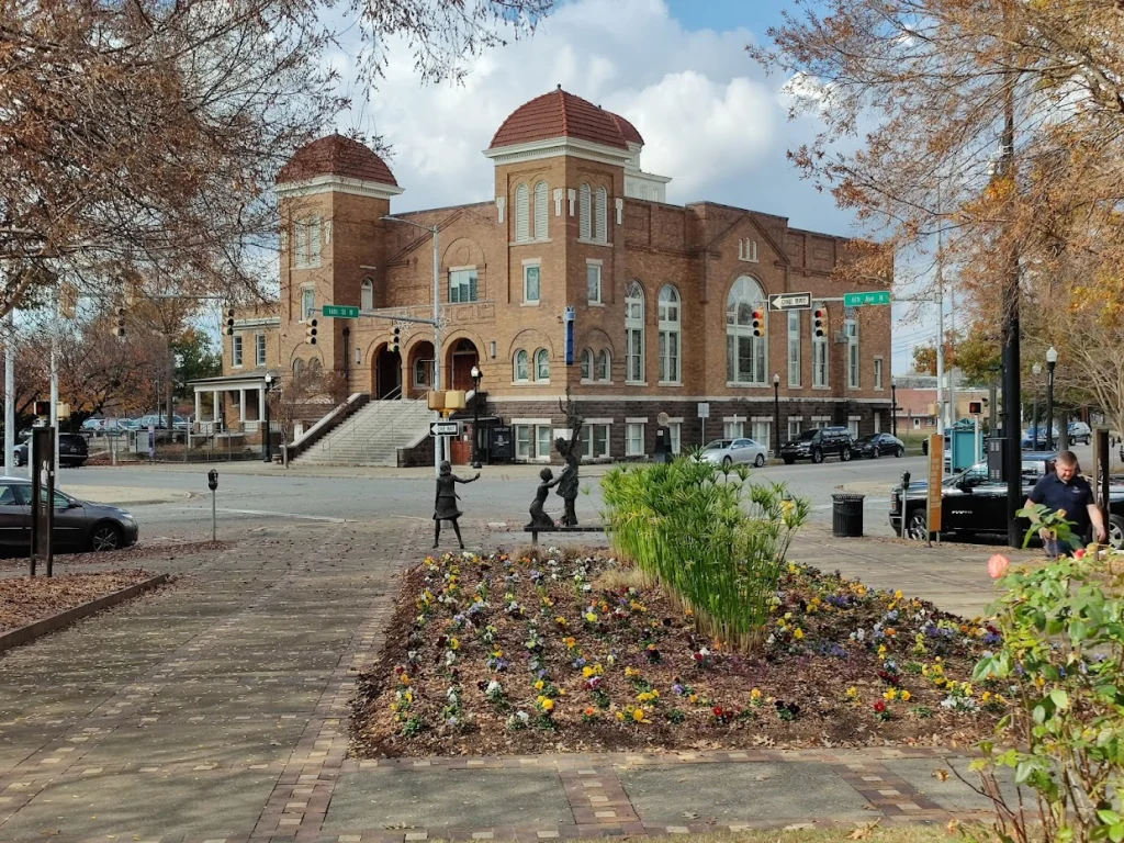 Straatbeeld in Birmingham met een bakstenen kerk met twee torens, bronzen kinderstandbeelden en een bloemperk op de voorgrond