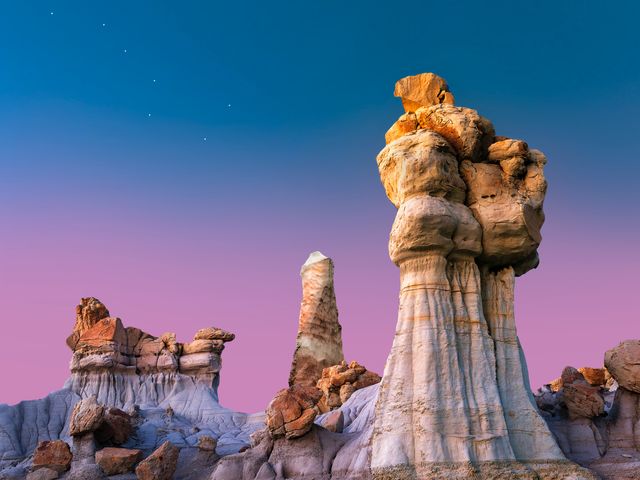 Grillig gevormde hoodoo rotszuilen van gelaagd gesteente steken af tegen een paarsblauwe avondlucht met sterren in de Bisti Badlands
