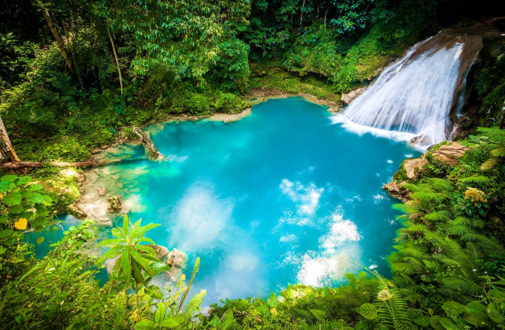 Uitzicht van bovenaf op een turkooizen poel met een waterval aan de zijkant omringd door groene jungle bij Blue Hole in Jamaica