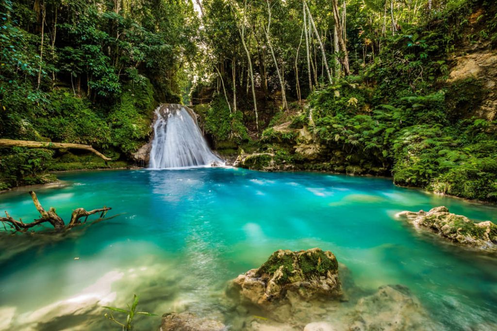 Een smalle waterval stroomt in een turkooizen poel tussen rotsen en dichte jungle bij Blue Hole in Jamaica