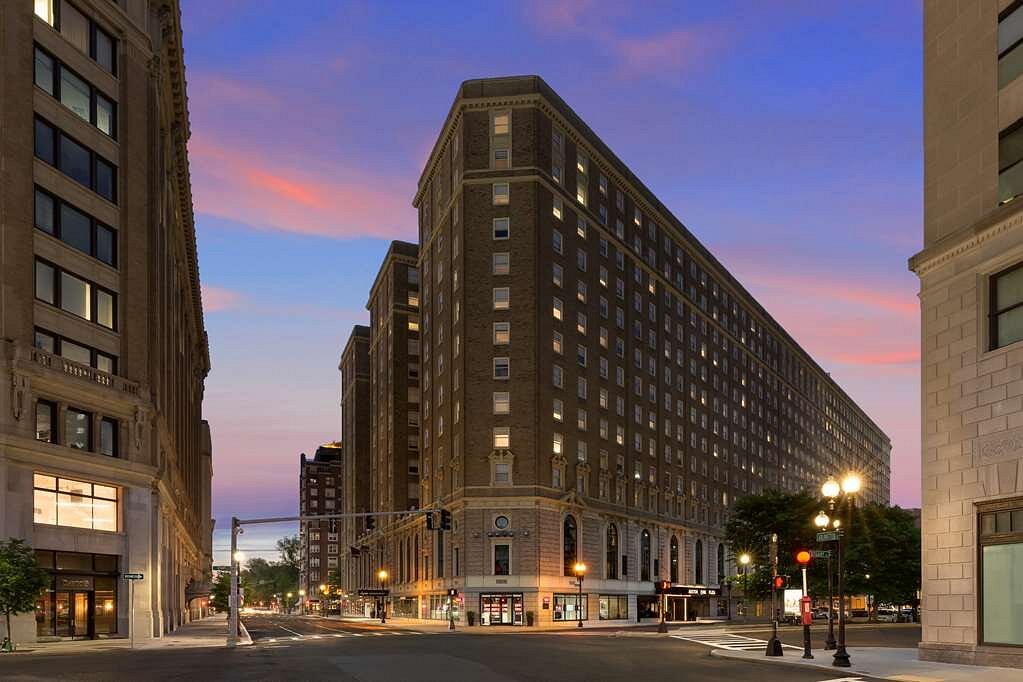 Hoog bakstenen hotelgebouw op een straathoek met verlichte ramen tegen een roze en blauwe avondlucht in Boston
