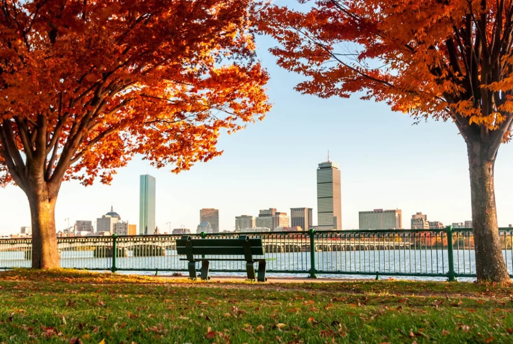 Rode herfstbomen omlijsten een bankje aan het water met de skyline van Boston en de Prudential toren op de achtergrond