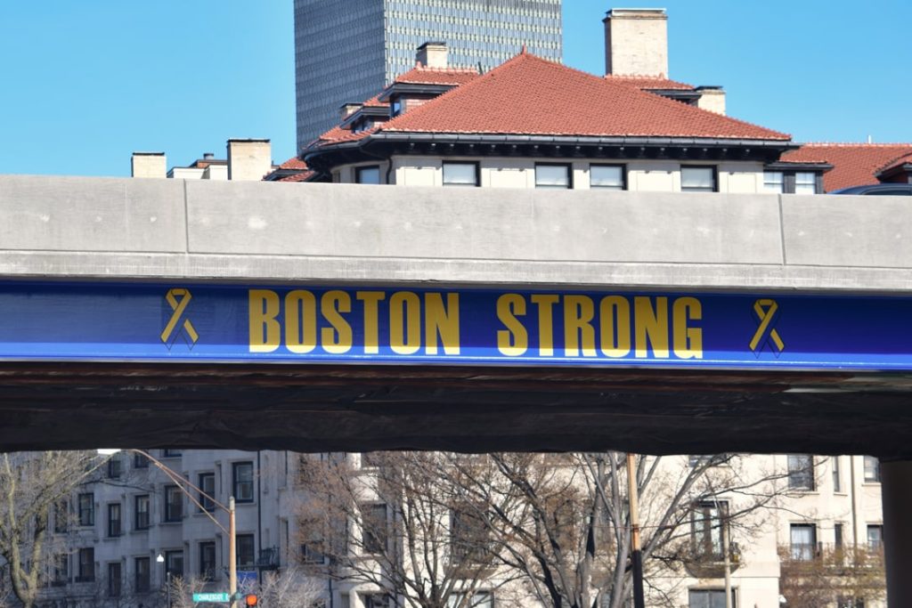 Blauw bord met gele letters Boston Strong en lintjes bevestigd aan een betonnen snelwegviaduct met gebouwen op de achtergrond