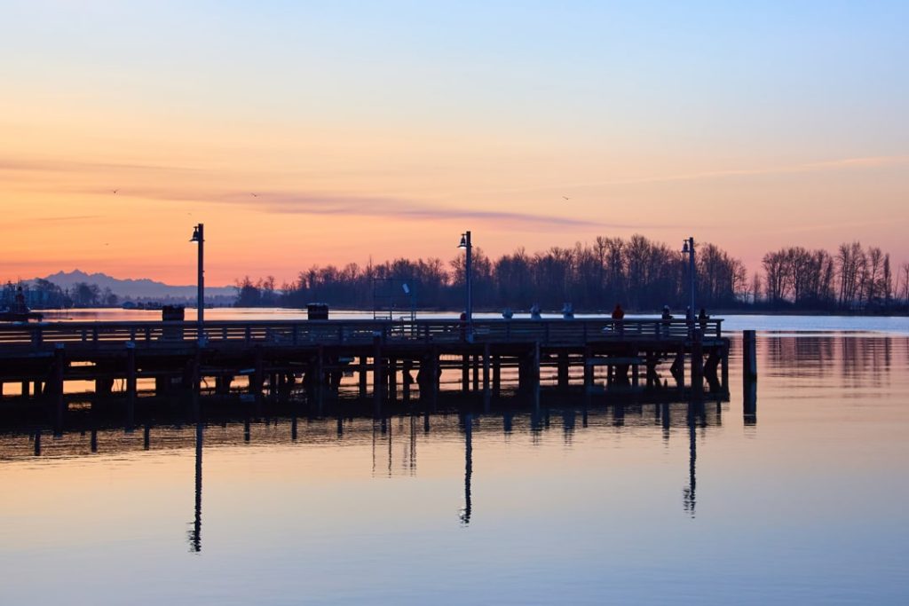 Lange houten steiger met lantaarnpalen, weerspiegeld in kalm water bij een oranje zonsondergang met bomen aan de horizon