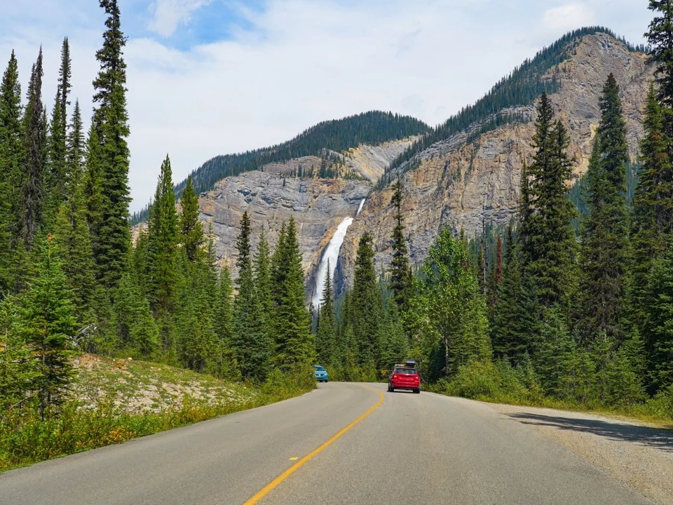 Een landweg omzoomd door dennenbomen leidt naar een hoge waterval tussen de bergen van British Columbia