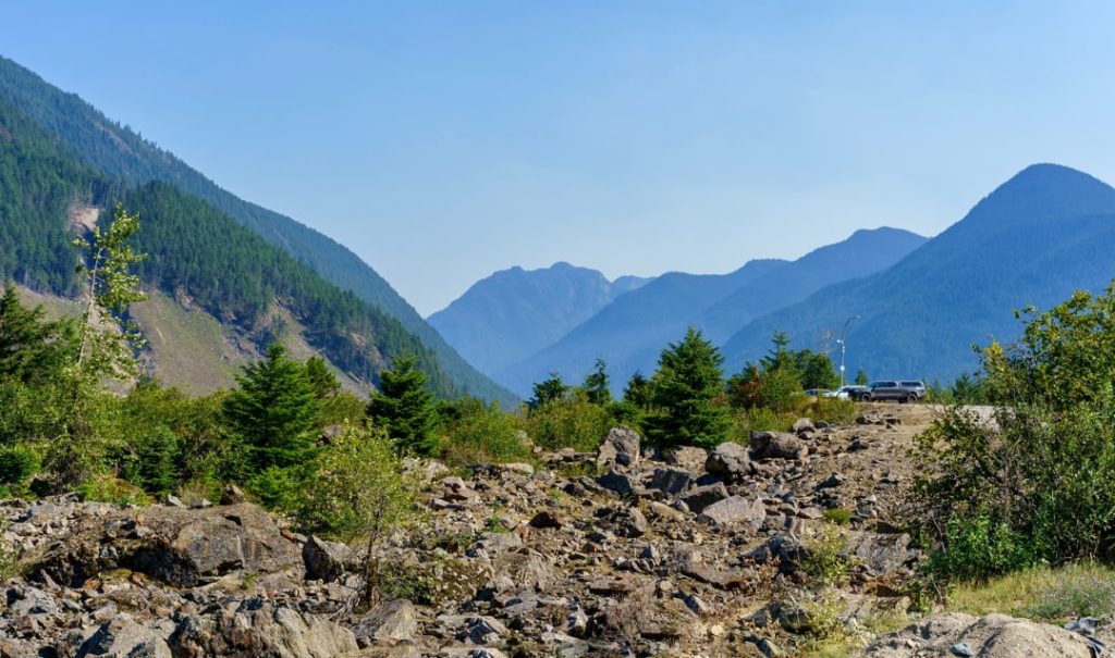 Verspreide rotsblokken liggen in een begroeid dal met dennenbomen terwijl beboste bergtoppen zich achter elkaar aftekenen in de verte