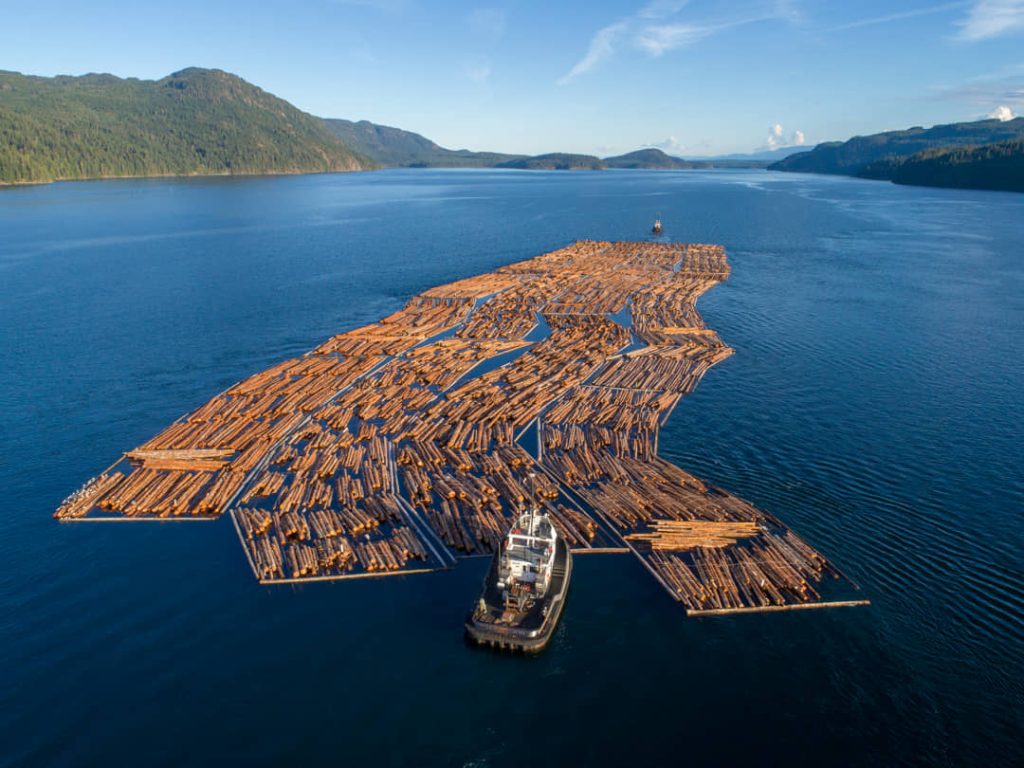 Luchtfoto van een sleepboot die een groot vlot boomstammen voorttrekt over kalm water tussen beboste heuvels