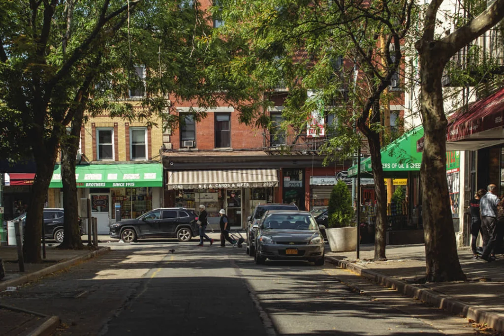 Rustige, boomrijke straat in de wijk Belmont in de Bronx met winkelpuien, geparkeerde auto's en voetgangers in de zon