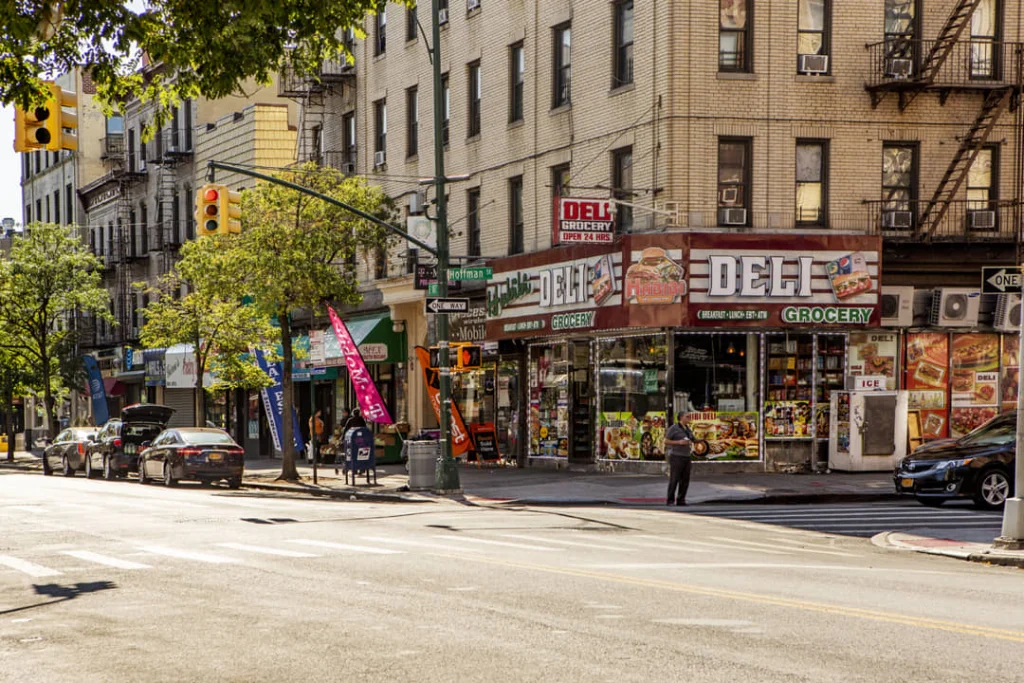 Zonnige straathoek in de wijk Belmont in de Bronx met een deli grocery winkel, geparkeerde auto's en bakstenen woongebouwen