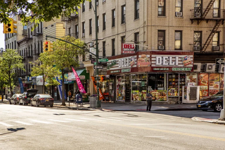 Zonnige straathoek in de wijk Belmont in de Bronx met een deli grocery winkel, geparkeerde auto's en bakstenen woongebouwen