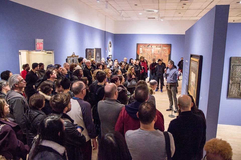 Grote groep museumbezoekers luistert naar een gids in een blauwe zaal met kunstwerken aan de muren in de Bronx