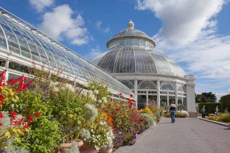 De koepelvormige glazen kas van de New York Botanical Garden omringd door kleurrijke bloemperken onder een blauwe lucht