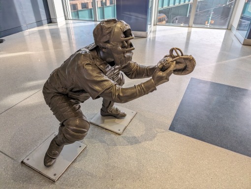 Levensgroot bronzen beeld van een honkbalvanger in hurkhouding met handschoen en helm op een museumvloer