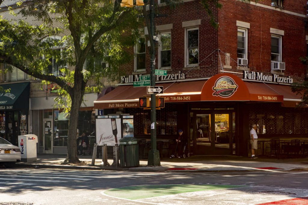 Bakstenen hoekpand met een rode luifel van een pizzeria en straatnaambordjes in de Italiaanse wijk Belmont in de Bronx