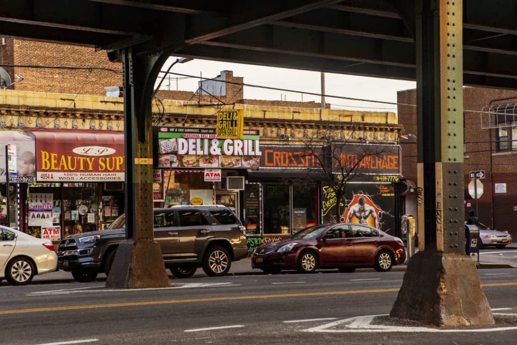 Winkels en auto's onder een verhoogd metroviaduct in Woodlawn in de Bronx met neonreclames van een deli en grillrestaurant
