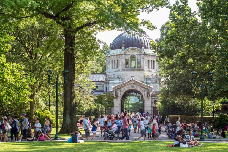 Bezoekers lopen door een groen park naar het monumentale koepelgebouw bij de ingang van de Bronx Zoo