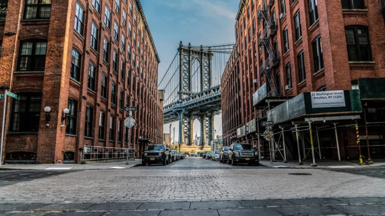 Kasseienstraat tussen bakstenen pakhuizen in Dumbo, Brooklyn met geparkeerde auto's en de Manhattan Bridge zichtbaar tussen de gebouwen