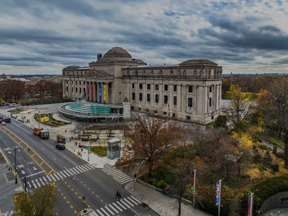 Luchtfoto van het neoclassieke gebouw van het Brooklyn Museum met koepeldak, brede trap en omliggend park onder een bewolkte lucht