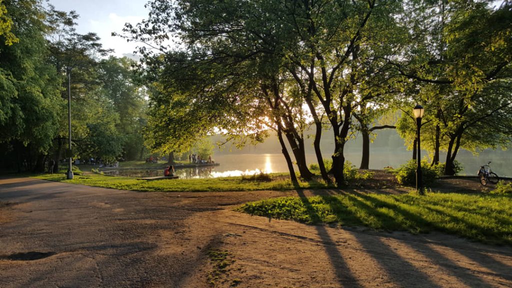Zandpad tussen bomen langs een vijver waar de avondzon doorheen schijnt, met mensen bij het water in Prospect Park in Brooklyn