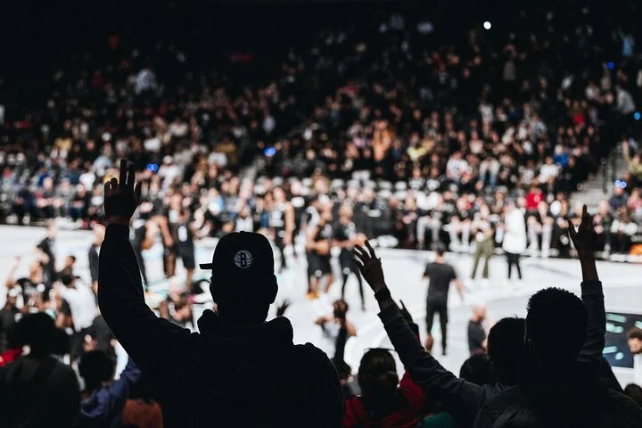 Toeschouwers steken hun handen omhoog tijdens een wedstrijd, op de voorgrond een bezoeker met een pet van de Brooklyn Nets
