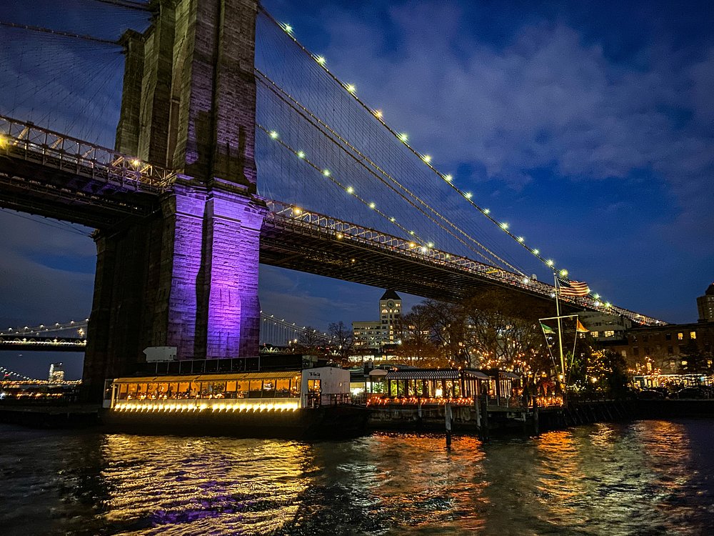 Verlicht restaurantschip River Cafe aan het water onder de paars verlichte pijler van de Brooklyn Bridge bij nacht