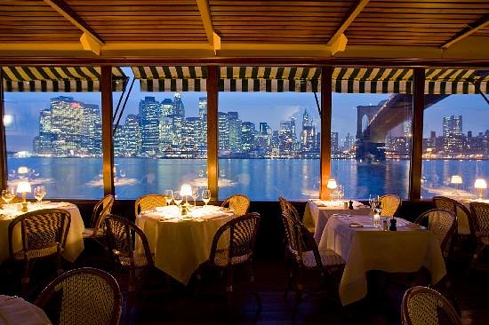 Gedekte tafels bij grote ramen met uitzicht op de verlichte skyline van Manhattan en de Brooklyn Bridge in het River Cafe