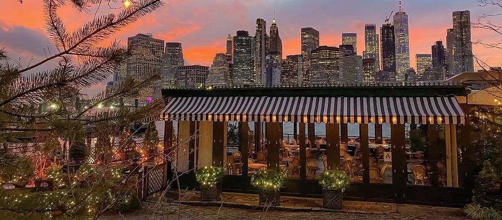 Overdekt terras met gestreepte luifel voor de skyline van Manhattan bij een felrode zonsondergang, gezien vanaf het River Cafe in Brooklyn