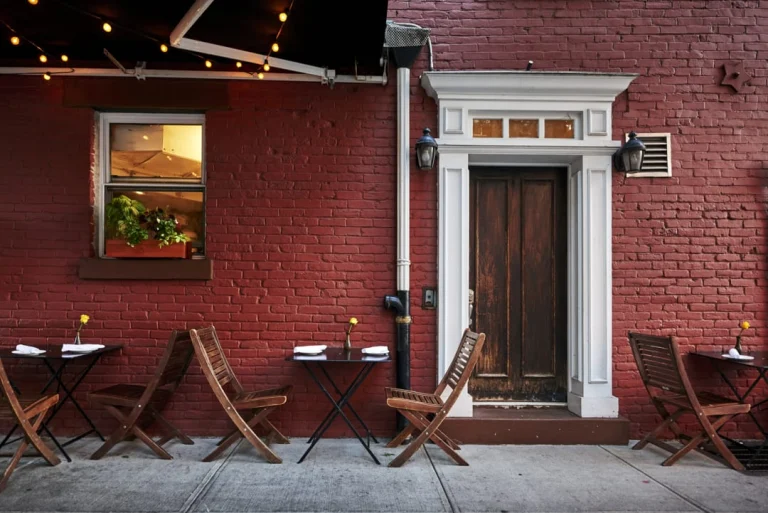 Houten terrastafels en stoelen voor een rode bakstenen gevel met een houten voordeur en lichtjes onder een luifel in Brooklyn