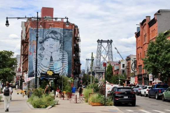 Levendige straat in Williamsburg in Brooklyn met een grote muurschildering van een kind op een gebouw en de Williamsburg Bridge op de achtergrond