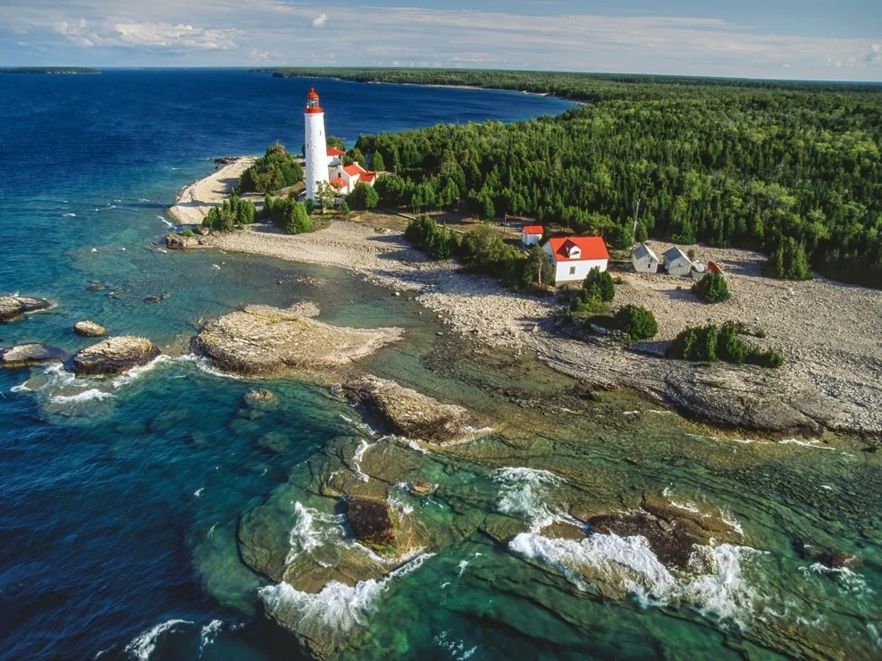 Witte vuurtoren met rode top op een rotsachtige landtong aan het heldere turquoise water van Bruce Peninsula, omringd door bos
