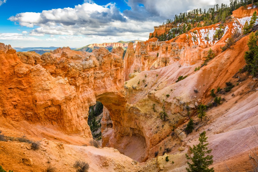 Kleine natuurlijke rotsboog gevormd tussen oranje pilaren, met dennenbomen op de rand van het amfitheater in Bryce Canyon