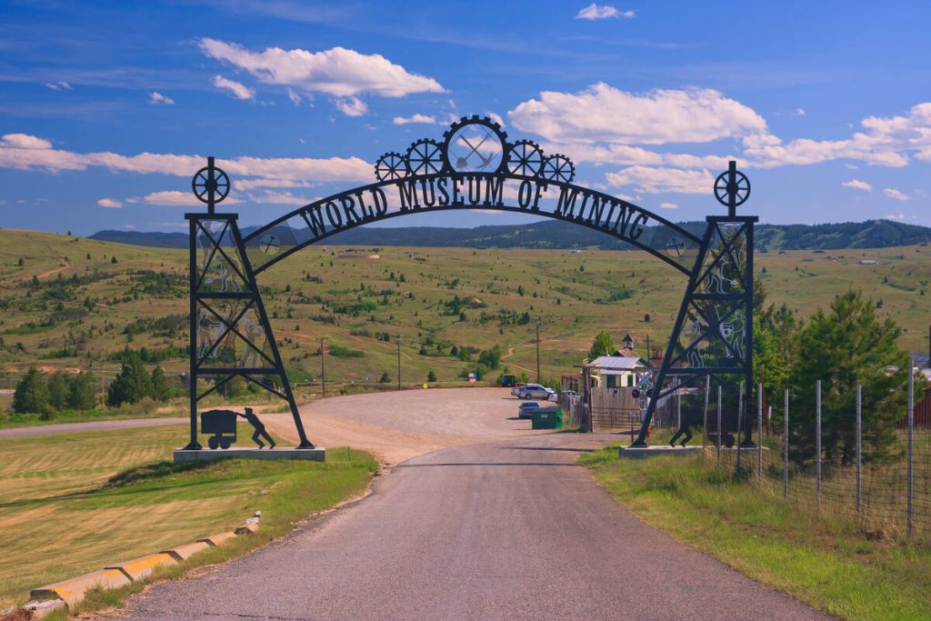 Metalen toegangspoort met tandwielen en de tekst World Museum of Mining boven een oprijlaan naar het openluchtmuseum in Butte