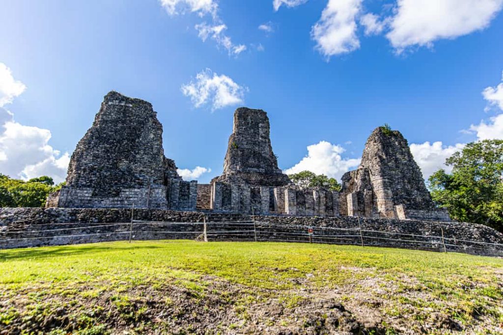 Drie verweerde stenen tempeltorens staan op een verhoogd platform met een grasveld ervoor bij Calakmul