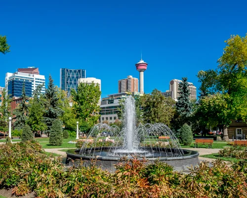 Klassieke fontein met opspattend water in een groen park met struiken op de voorgrond en de Calgary Tower tussen kantoorgebouwen op de achtergrond