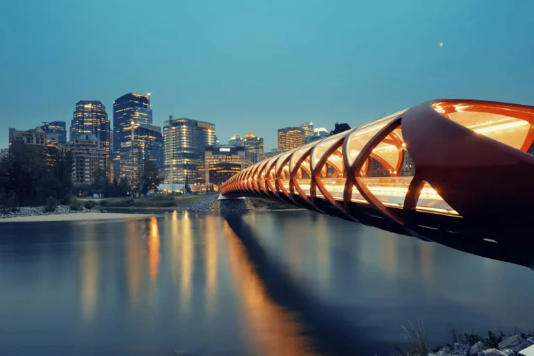 De rood verlichte Peace Bridge in Calgary weerspiegelt in de rivier terwijl op de achtergrond de skyline van de stad oplicht in de schemering