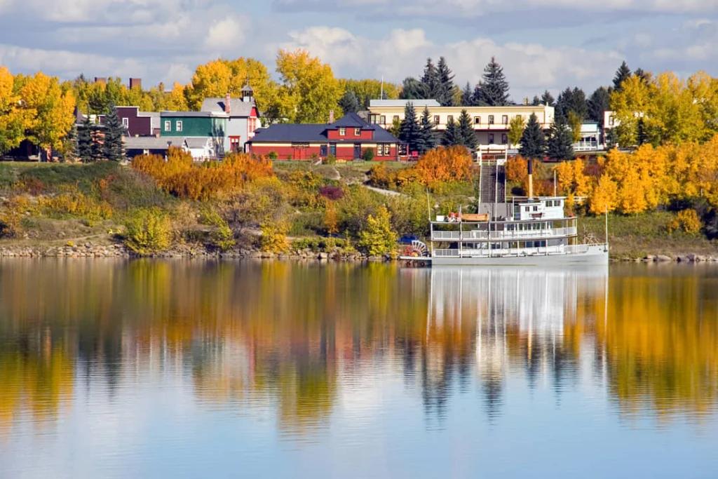 Een witte historische raderboot ligt aangemeerd op een rivier bij Calgary, met een dorpje en bomen in herfstkleuren op de achtergrond weerspiegeld in het water