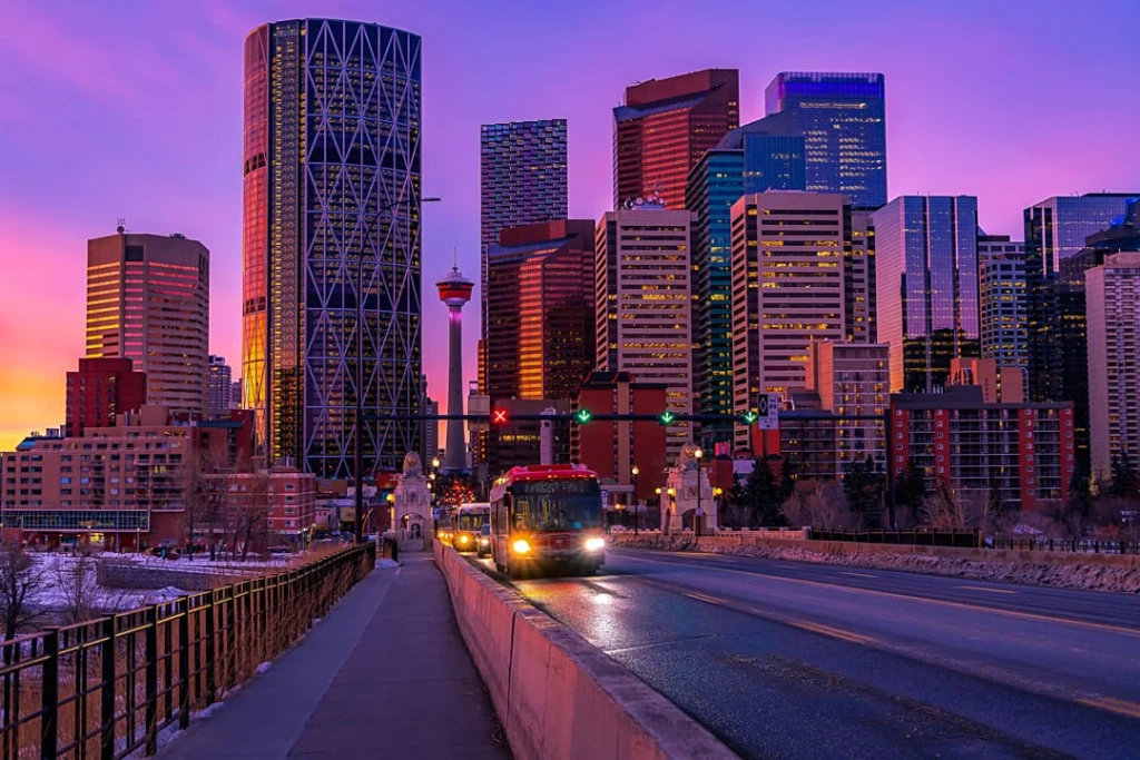 Wolkenkrabbers en de Calgary Tower steken af tegen een paarsroze avondlucht terwijl bussen over de brug rijden