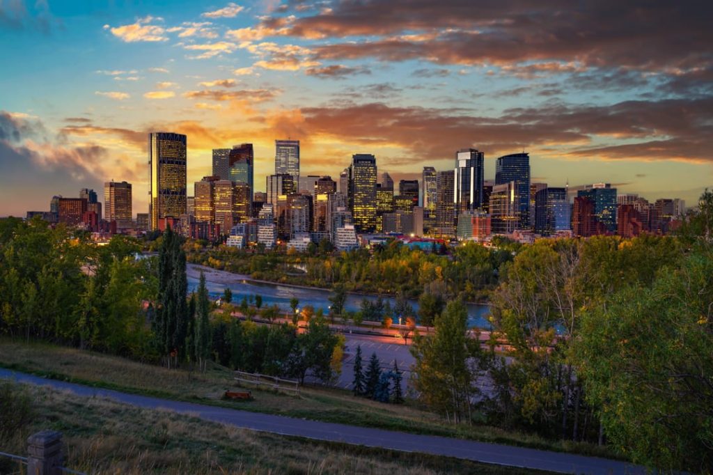 De skyline van Calgary met verlichte wolkenkrabbers weerspiegelt tegen een oranje avondlucht boven de rivier en groene parken