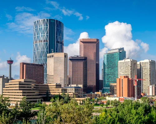 Moderne wolkenkrabbers van verschillende vormen en kleuren met de Calgary Tower op de achtergrond en groene bomen op de voorgrond onder een blauwe lucht
