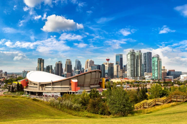 Uitzicht over Calgary met de wolkenkrabbers en het ronde Saddledome-stadion, omgeven door groene heuvels en bomen onder een blauwe lucht