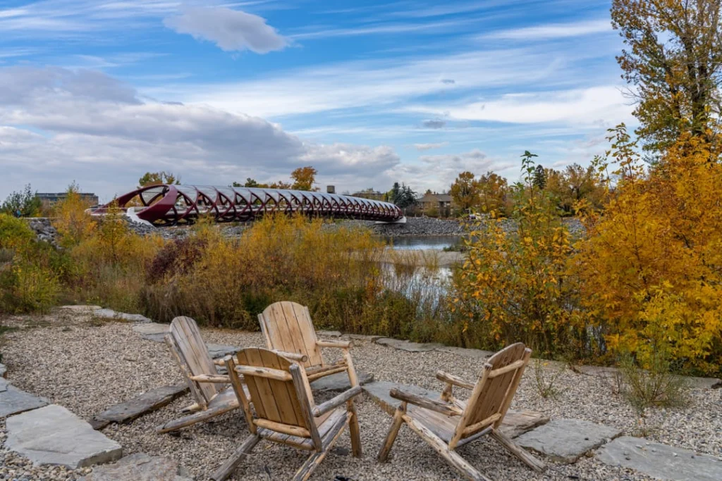 Houten tuinstoelen op een grindpad met uitzicht op de rode Peace Bridge en herfstbomen in Prince's Island Park