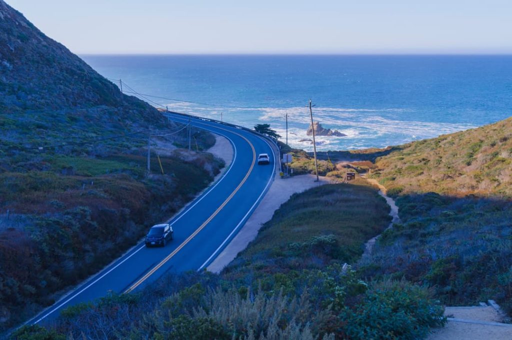 Lege kustweg die langs struikgewas en een klif slingert, met auto's in de verte en de oceaan die zich uitstrekt tot de horizon in het avondlicht