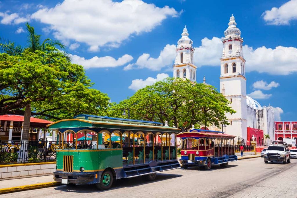 Twee kleurrijke trolleybussen staan geparkeerd voor de witte kathedraal met twee torens in het centrum van Campeche